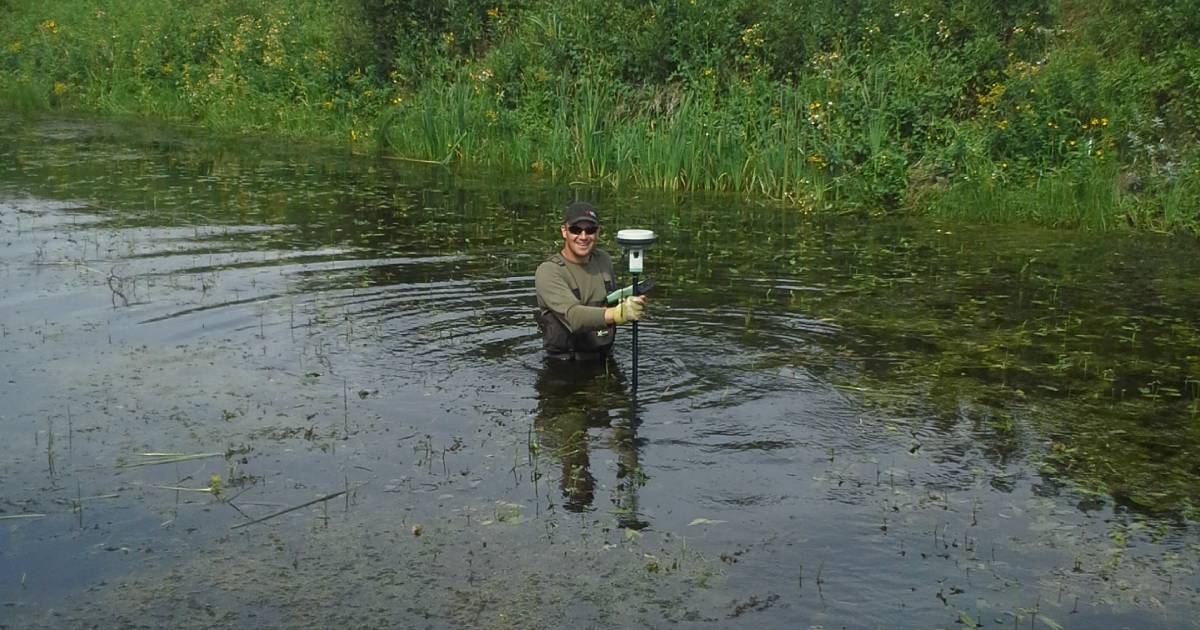 Surveyor wading in a river with a GPS rod conducting traditional bathymetric measurements — exposing crew to current, footing risk, and wildlife